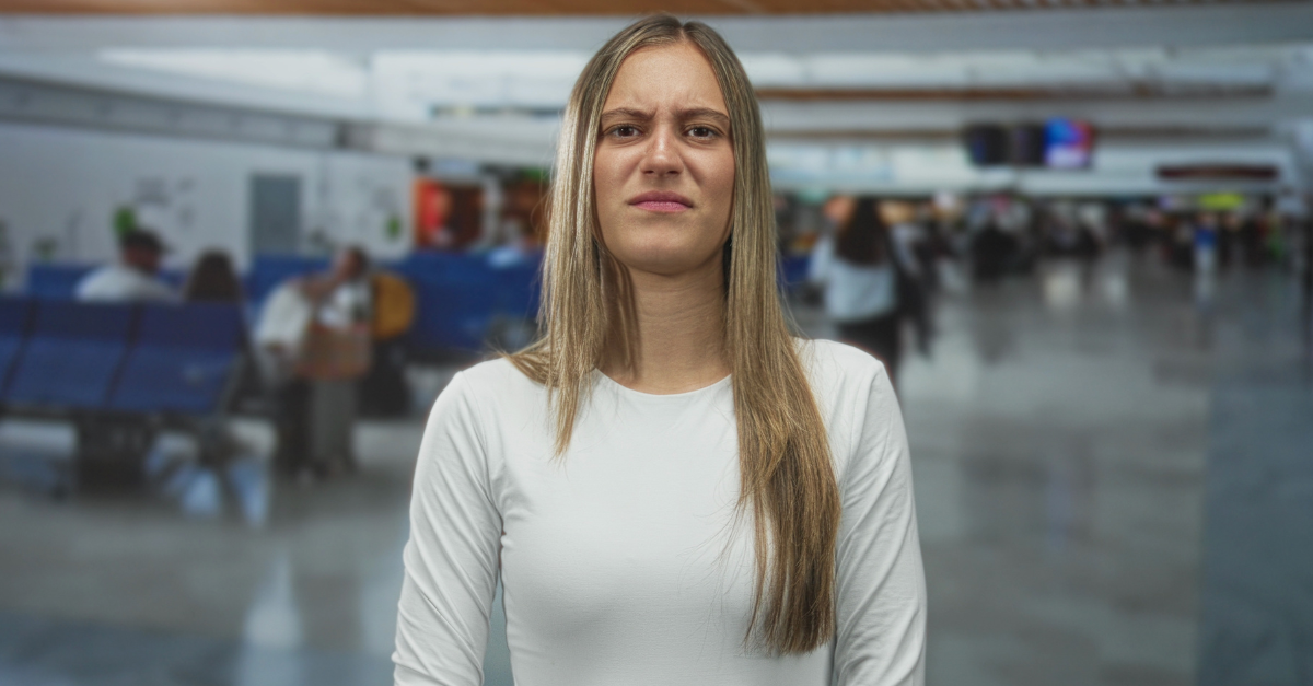 Woman frowning with scrunched face and white long sleeve top at airport terminal; annoyance travel stress.