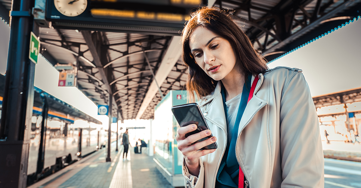 Young woman with departure times behind her waiting for her train while holding her mobile phone - Woman looking at the clock in the train station while her train is delayed - Transportation concept
