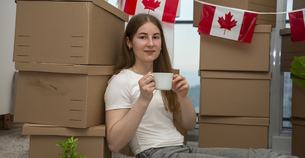 A young woman sits among stacked moving boxes while holding a coffee cup, with Canadian flags display