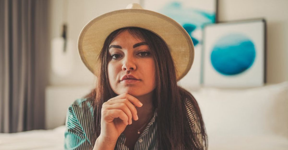 Woman with sun hat resting on bed in stylish indoor setting with modern art decor.