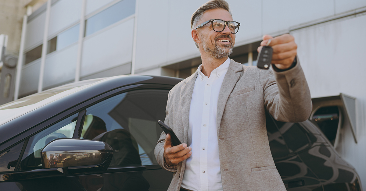 Adult man customer male buyer client wearing white shirt classic grey suit standing near black electric car while charging looking aside use mobile cell phone hold give keys outside. Business concept