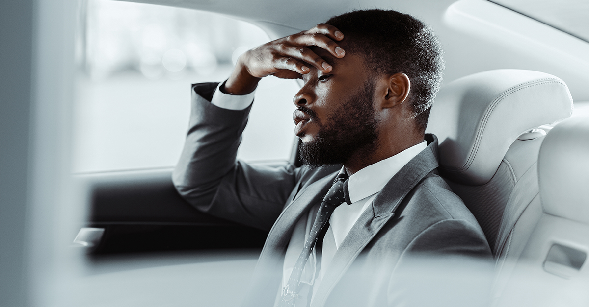 A Black man in a grey suit sits in the back seat of a car. He looks down and has his hand on his forehead as if he is exhausted or stressed. The image is taken from inside the car