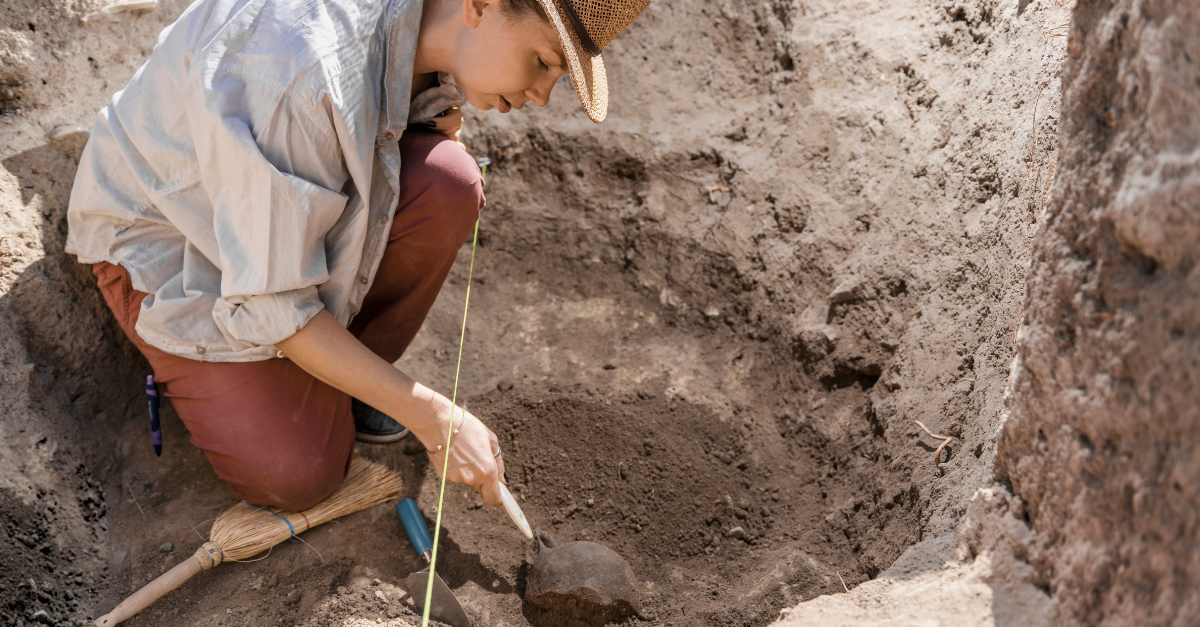 Archaeologist digging with hand trowel, recovering ancient pottery object from an archaeological site.