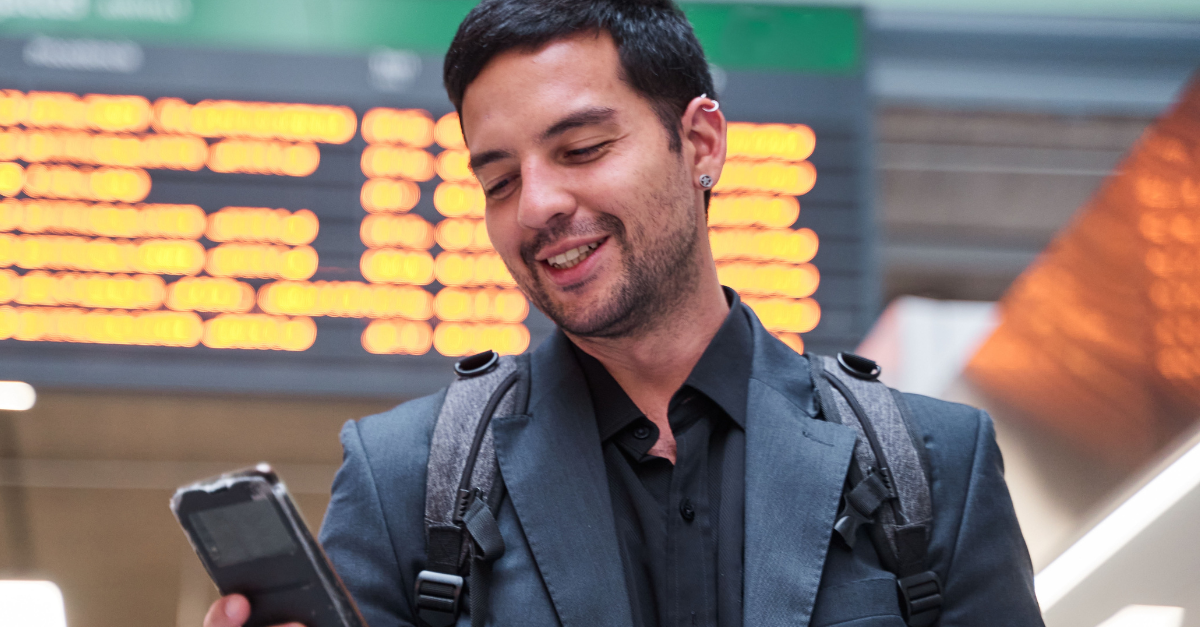Young latino man travelling the world. man looking at his smart phone in the station with the travel schedule in the background.