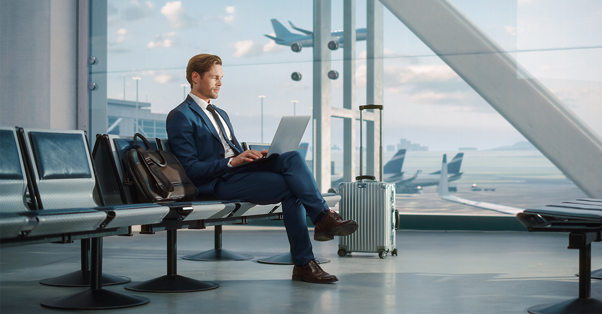 Modern Airport Terminal: Handsome Businessman Working on Laptop Computer While Waiting for His Flight. Man Sitting in a Boarding Lounge of Big Airline Hub with Airplanes Departing and Arriving
